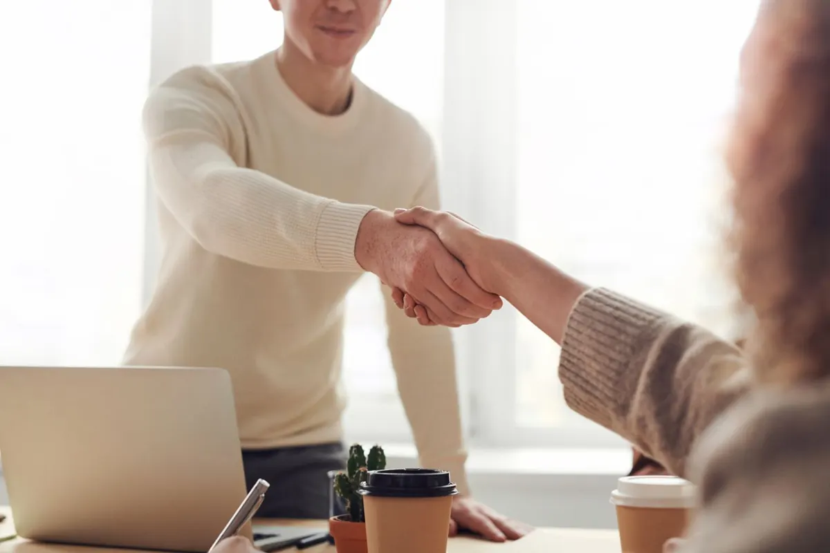 Two people shaking hands over a desk with a laptop